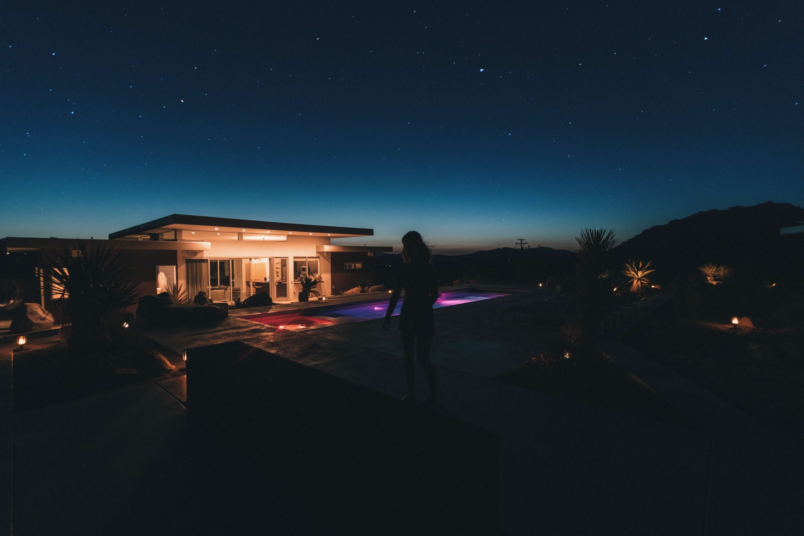 Desert backyard landscape at dusk with modern home, illuminated pool, and starry sky, showcasing Arizona's unique outdoor living and landscaping trends.