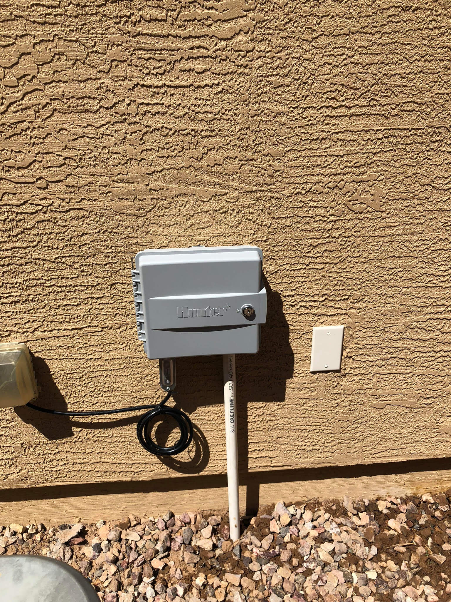 Hunter Hydrawise WiFi irrigation controller mounted on a textured wall, connected to a water line, showcasing professional installation by Core Landscape in Phoenix, AZ.