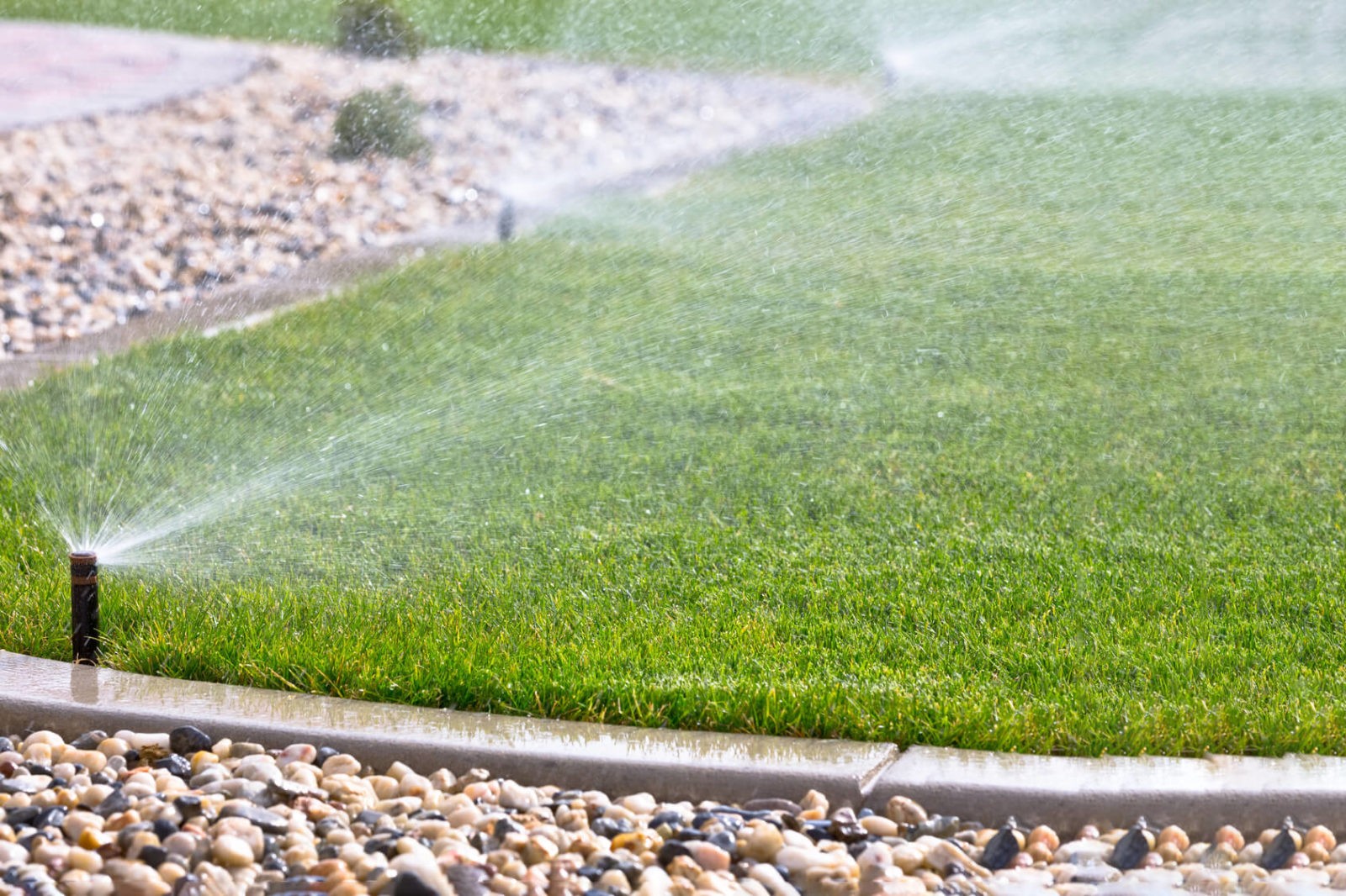 Lawn sprinkler system watering green grass in landscaped yard with pebbled border, illustrating irrigation efficiency for homeowners in Phoenix and central Arizona.